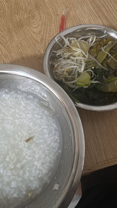 A stainless steel bowl of white rice congee sits next to a side dish of bean sprouts and dark leafy greens on a wooden table.