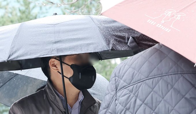 A photo of a person wearing a black face mask standing under a black umbrella in the rain, next to another person holding a maroon umbrella.