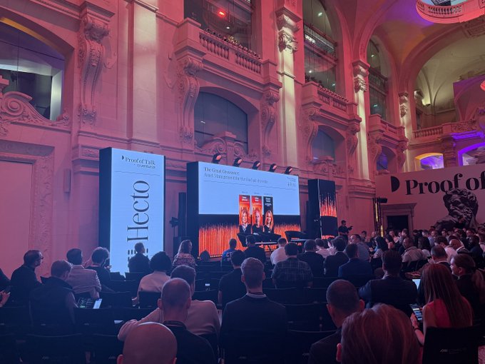 The image shows a large room with a stage set up for an event, featuring two speakers standing behind a screen displaying their names and titles. The audience consists of people sitting in rows of chairs facing the stage. A sign reading "Profo" is visible on one side of the room.