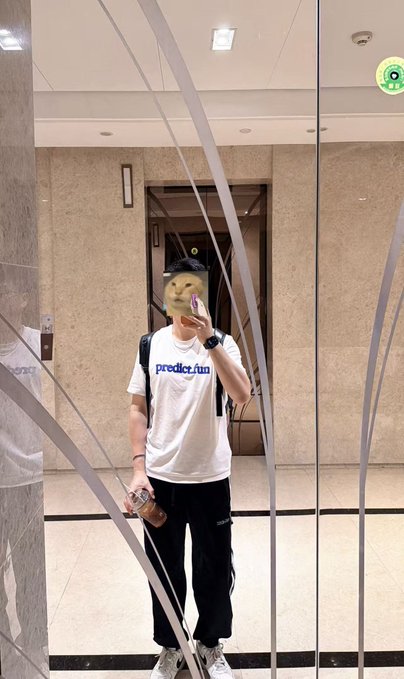 A person takes a mirror selfie in an elevator wearing a white 'predict.fun' t-shirt, black pants, and holding a coffee cup, with their face obscured by a sticker.