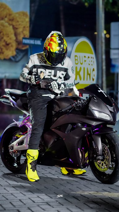 A motorcyclist wearing a yellow and black helmet and white 'DEUS' jacket sitting on a black sportbike, with a 'BURGER KING' sign visible in the background.