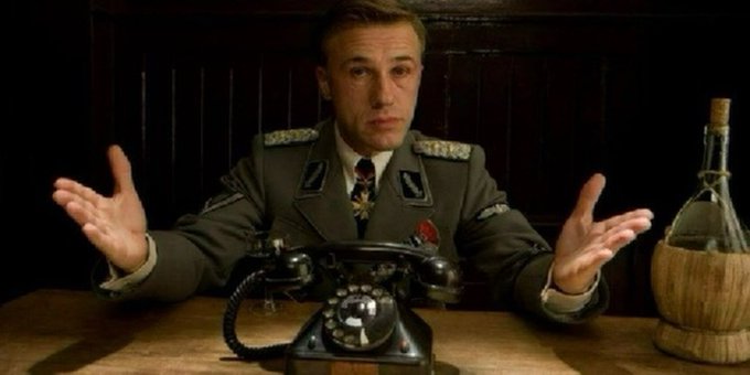 A stern man in a Nazi-style uniform sits at a desk with palms raised in a questioning gesture, a vintage rotary telephone before him and a straw-wrapped bottle to his side. The image humorously evokes a bureaucratic, authoritarian tone—fitting the post's quip about an absurd, overly ambitious plan, as if interrogating the logic of "building a launchpad for launchpads."