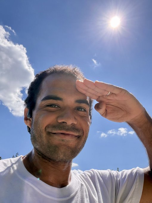 A man takes a selfie outdoors, shielding his eyes from the bright sun with his hand while smiling against a vivid blue sky. The sun-drenched image mirrors the post's energetic vibe of completing a 2-mile walk, with the sunny backdrop symbolizing the warmth and positivity behind his mention of Arbitrum and Ethereum.