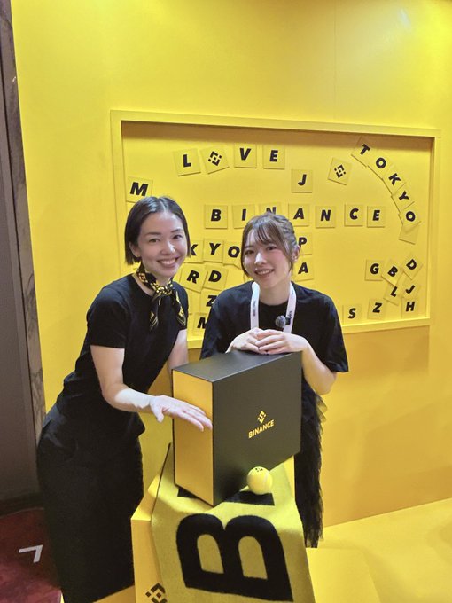 Two smiling women pose together at a Binance-branded booth, with one wearing a yellow "BINANCE" lanyard and the other sporting a yellow patterned scarf, standing beside a yellow box displaying the Binance logo. Behind them, a yellow wall features Scrabble-like tiles spelling out "BINANCE," "TOKYO," and "LOVE," along with other scattered letters, capturing a cheerful moment at what appears to be a Binance event in Tokyo.