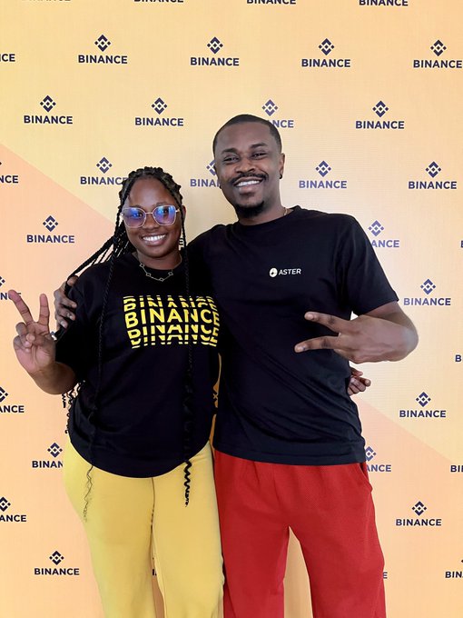 Two people posing in front of a Binance branded backdrop; the woman wears a 'BINANCE' t-shirt and yellow pants, while the man wears an 'ASTER' t-shirt and red pants.