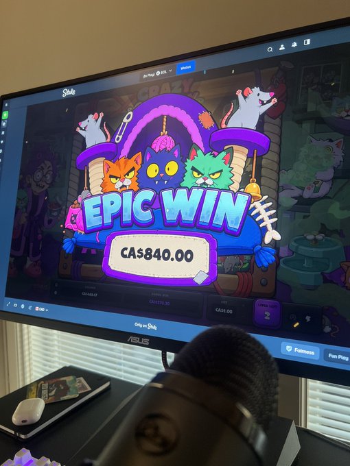 An ASUS computer monitor displaying an 'EPIC WIN' of CA$40.00 on a cat-themed online slot game called 'The Crazy Ones' by Sloto, with a microphone and phone visible on the desk.