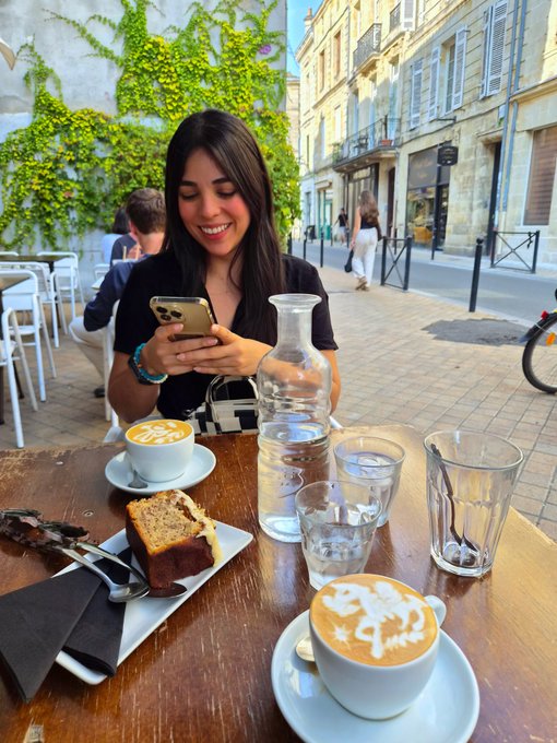 A young woman sits at an outdoor cafe table smiling at her phone, with two cappuccinos featuring latte art, a slice of cake, a glass water carafe, and empty glasses on the table, set against a charming European street backdrop with ivy-covered walls.