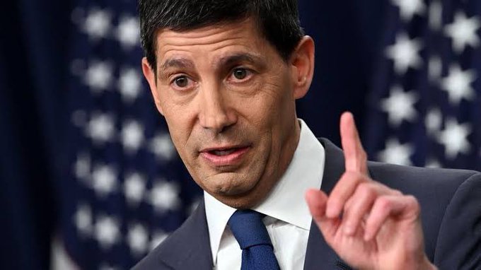 A businessman in a suit speaks while gesturing with his finger raised, set against a backdrop of American flags. The image likely accompanies the post about upcoming market-moving economic events, suggesting commentary on U.S. financial data releases.