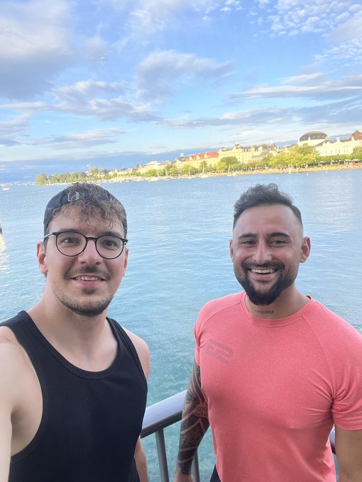 Two friends smile for a selfie on the shore of Lake Zurich, with the calm blue water and distant cityscape in the background, perfectly capturing the relaxed "gm" vibe of a fresh new week.