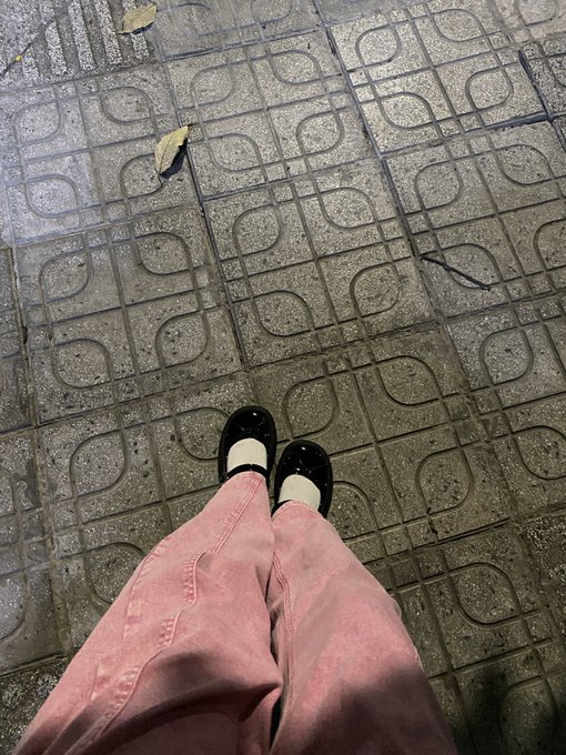 A first-person view looking down at someone wearing pink pants, white socks, and black Mary Jane shoes standing on patterned tile pavement at night.