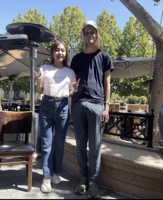A young woman and a young man pose for a photo outdoors at what appears to be a cafe patio, with the woman making peace signs.