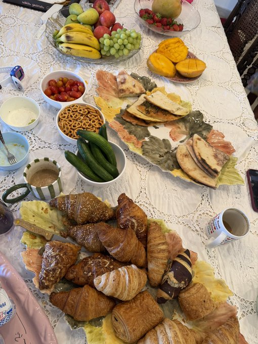 A table filled with an assortment of foods including croissants, fresh fruit like bananas, apples, grapes, and mangoes, along with vegetables, dips, and crackers arranged on a white tablecloth.