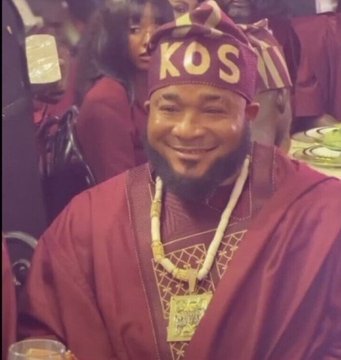 A smiling man wearing traditional maroon attire and a cap with 'KOS' written on it, adorned with a gold necklace.
