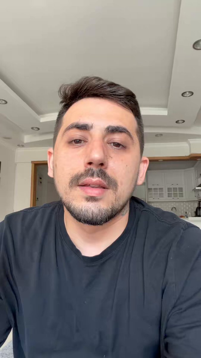 The image features a man wearing a black shirt with his face partially visible as he looks directly at the camera. The background of the image shows a kitchen area with white cabinets, suggesting that this photo was taken in someone's home.