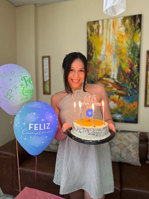 A smiling woman in a striped halter dress holds a birthday cake with lit candles, standing next to balloons that say 'Feliz Cumpleaños.'