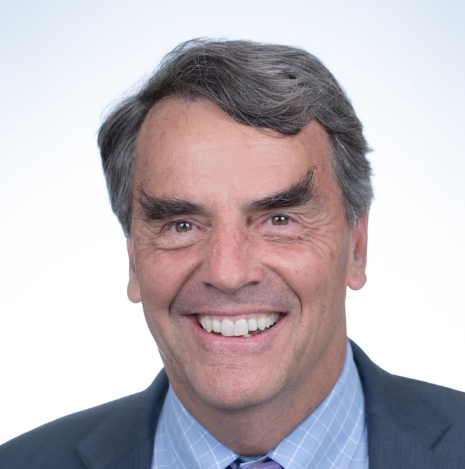 A headshot of a smiling middle-aged man with graying hair wearing a dark suit and light blue checkered shirt against a white background. The image appears to represent Charles Hoskinson, the founder of Cardano, whose leadership of the $ADA project the caption references.