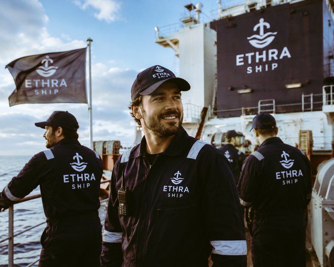 A confident sailor in a branded uniform smiles on the deck of a large ship, with his crew members working behind him. The matching gear, flag, and ship's hull all prominently display the "ETHRA SHIP" logo and anchor emblem, visually reinforcing the post's introduction of the @EthraShip project.