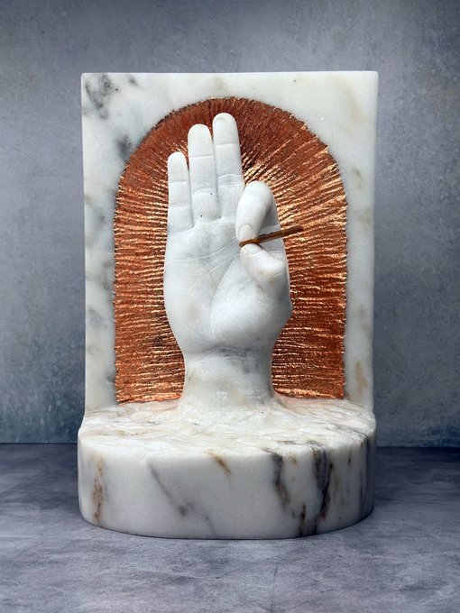 The image features a white marble statue of a hand holding a book, which appears to be a tribute to Bitcoiners. The hand is depicted in a "Gyan Mudra" or "Mudra of Wisdom," symbolizing knowledge and learning. The statue is placed on a gray surface against a grey background, creating a minimalist aesthetic that highlights the subject matter.