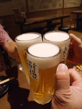 Three glasses of Japanese beer being clinked together in a toast at what appears to be an izakaya restaurant, with Japanese characters visible on the glasses.