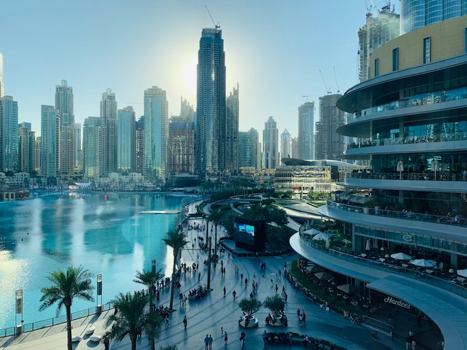 A bright, futuristic cityscape view of a waterfront promenade with modern skyscrapers, palm trees, a curved multi-level building, and crowds of people walking around a fountain.