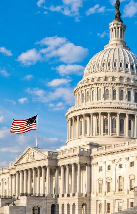 The United States Capitol building stands prominently beneath a partly cloudy blue sky, with the American flag flying beside its iconic dome and columned facade. The image of this legislative seat underscores the post's report that the CLARITY Act has stalled, as the House has canceled late September sessions, leaving minimal legislative days before the midterms.