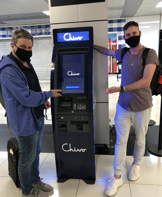 Two men wearing masks stand on either side of a "Chivo" branded Bitcoin ATM in an airport terminal, with one pointing at the screen and the other gesturing toward it, testing out the machine that became part of El Salvador's historic adoption of Bitcoin as legal tender on September 4, 2021.