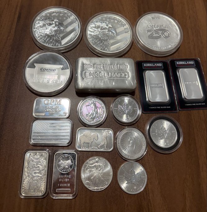 A collection of silver bullion items including American Silver Eagles, Engelhard, Costco, Kirkland, and OPM bars, along with various rounds and 1 oz silver pieces, laid out on a wooden surface—reflecting a personal silver investment stash and the post's warning to halt further purchasing.