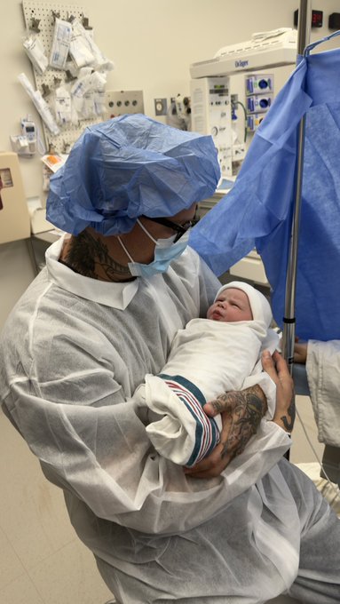 A man in surgical scrubs, cap, and mask cradles a swaddled newborn in a hospital delivery room.