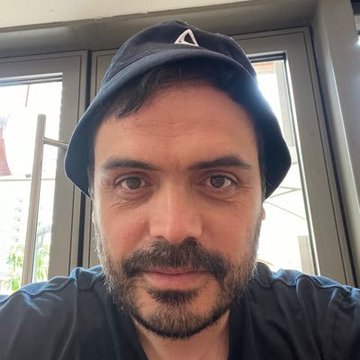 A selfie of a bearded man wearing a dark blue bucket hat, looking directly at the camera near a window.