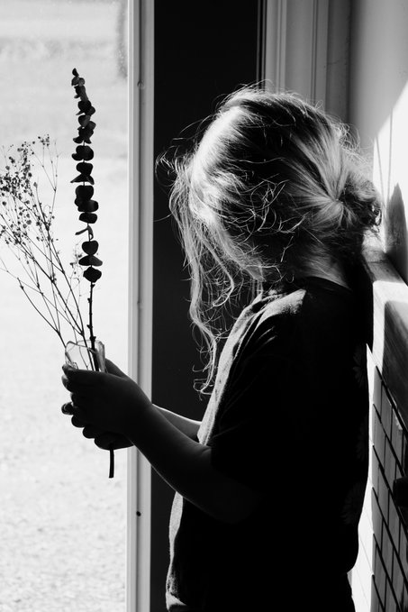 A black and white photograph of a person with long blonde hair holding dried flowers by a window, silhouetted against bright light.