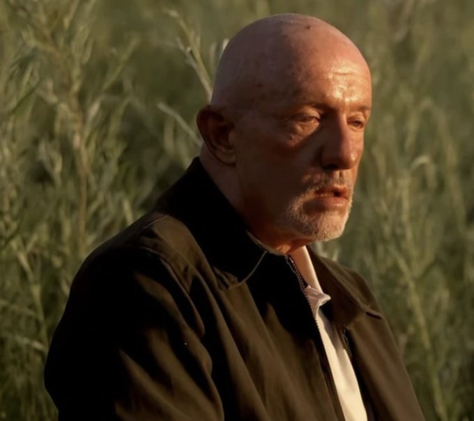 An older bald man with a white beard stands in a field of tall grass, wearing a black jacket over a white shirt, with a weary and resigned expression on his face. His dejected look visually captures the humor of the post, implying that despite years in crypto, he's far from wealthy and is instead left looking exhausted and disillusioned.