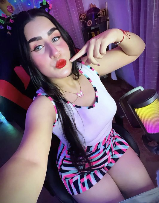 A woman with long dark hair poses seductively, wearing heavy makeup with red lipstick, a white top, and patterned shorts, sitting in what appears to be a gaming chair with colorful lighting in the background.