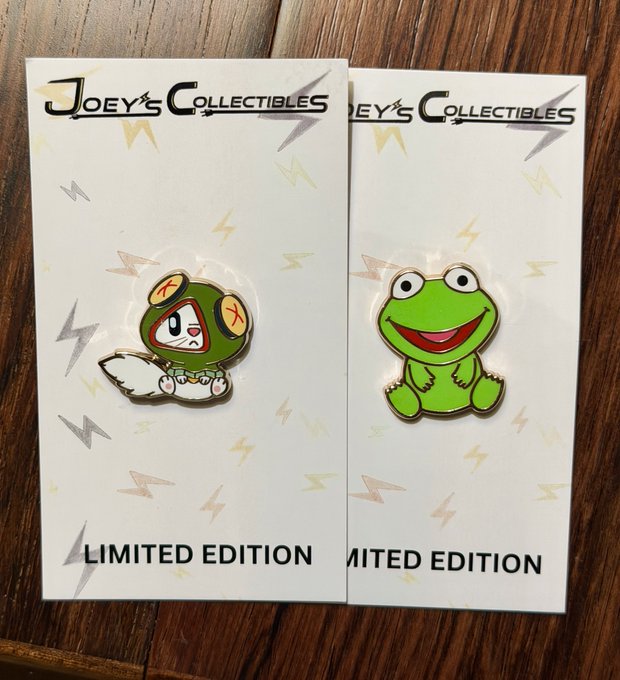 Two limited edition enamel pins from 'Joey's Collectibles' displayed on their packaging cards against a wooden surface, featuring a green hooded character and a smiling Kermit the Frog.