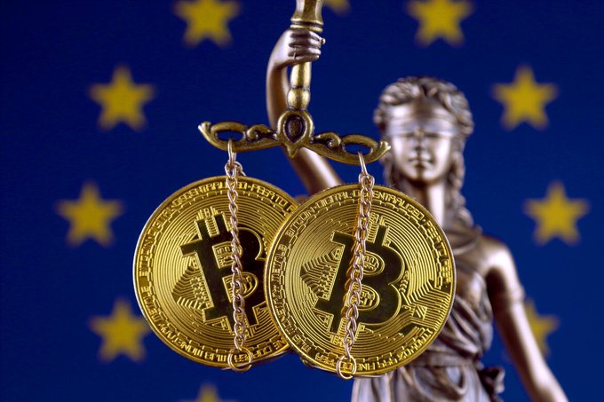 Two gold Bitcoin tokens hang from an ornamental scale held by Lady Justice, set against the European Union flag's yellow stars on blue, symbolizing the EU's regulatory scrutiny of cryptocurrency. The image visually reinforces the post's claim about EU lawmakers rushing to prepare "MiCA 2.0" stablecoin regulations in response to the U.S. GENIUS Act.