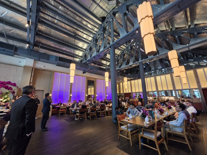An elegant restaurant interior with high industrial ceilings, hanging cylindrical lights, and diners seated at long tables while a man in a suit stands in the foreground.