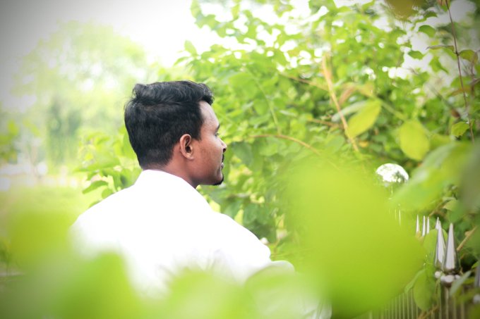 A young man in a white shirt seen in profile from behind, surrounded by lush green foliage with a metal fence partially visible.