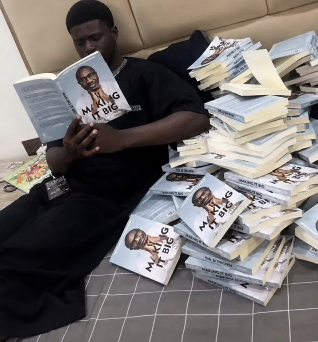 A young man in a black t-shirt sits on a bed reading a copy of the book 'Making It Big' by Max Inflictor, surrounded by a massive pile of identical copies.