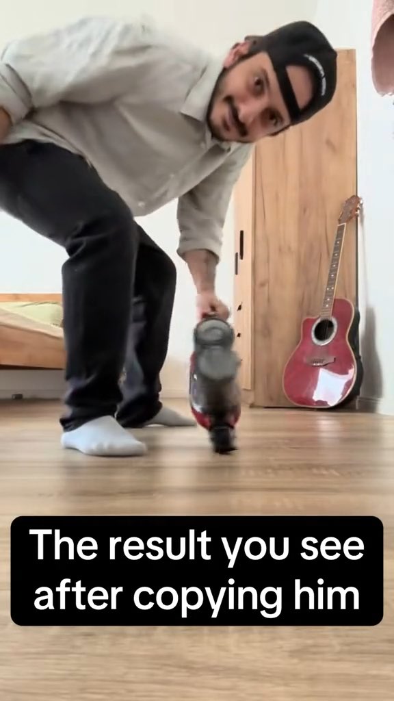A humorous video still of a man wearing a cap, shirt, and dark pants holding a jug in a room with a guitar, with caption 'The result you see after copying him'.