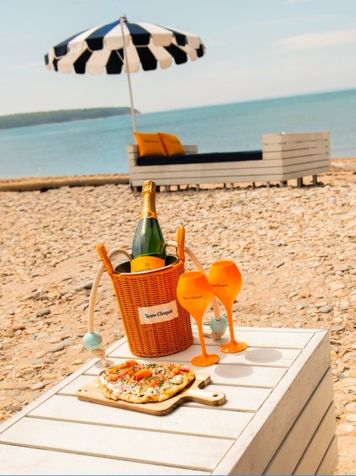 A relaxing beach setup features a bottle of Veuve Clicquot champagne chilling in an orange basket, two glasses, and a small pizza on a white wooden table, with a striped umbrella and lounge in the background.