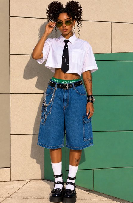 A fashion photo of a woman wearing a cropped white shirt, a black cross-pattern tie, denim jorts with chains, white socks, and black Mary Jane shoes, posing against a green and beige wall.