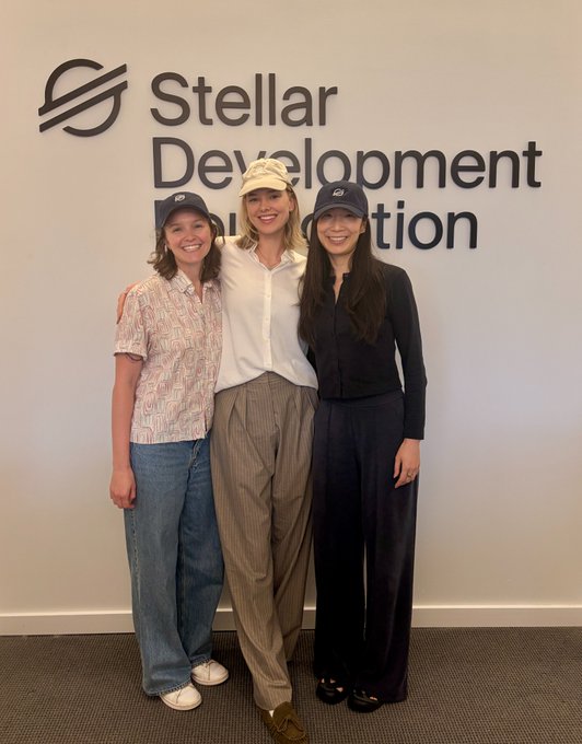 Three young women wearing baseball caps pose together in front of a wall with the 'Stellar Development' company logo, dressed in casual business attire.