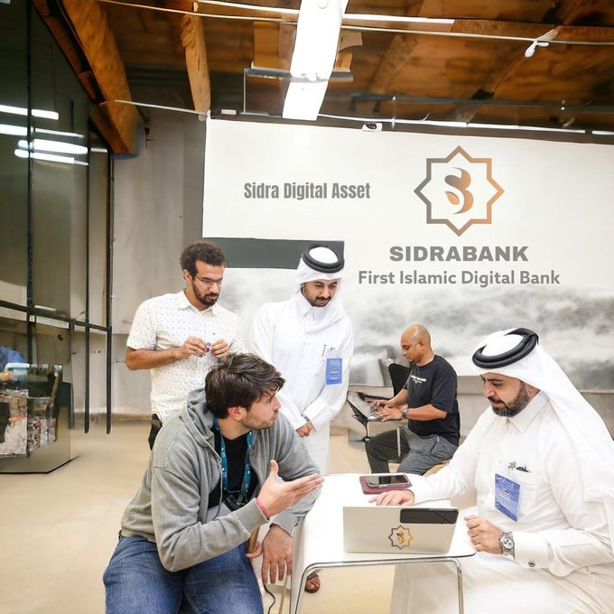 A photo taken at an event showing people interacting at a 'Sidra Digital Asset' booth for 'SIDRABANK First Islamic Digital Bank', with attendees in both Western and traditional Gulf attire.