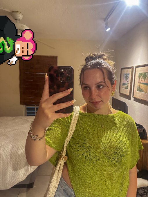 A mirror selfie of a woman in a green top holding a phone, with pixel art of a character with a pink hat and green hair in the upper left corner.