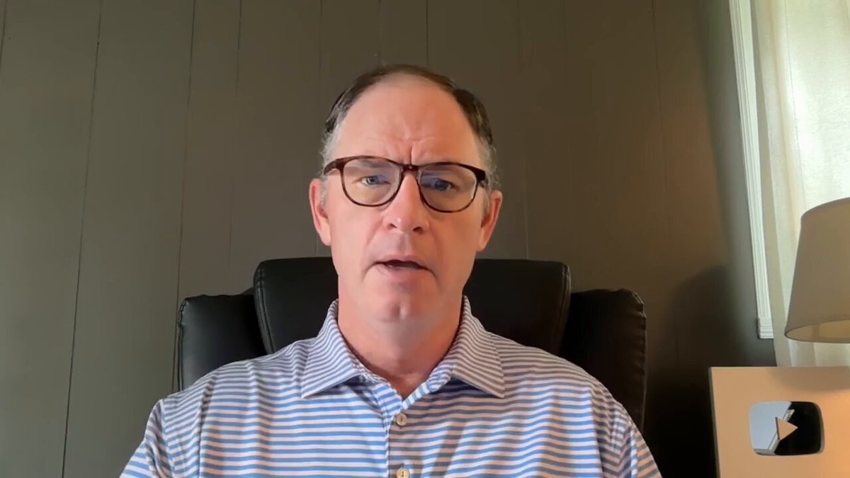 The image shows a man wearing glasses sitting in an office chair, looking at the camera with a serious expression. He appears to be giving a speech or presentation, as indicated by his posture and facial expressions. The background of the image features a gray wall, which complements the man's attire and adds depth to the scene.