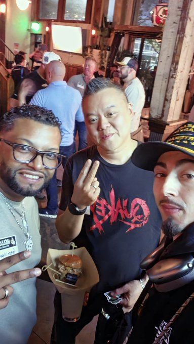 Three men pose for a selfie; one holds a small burger, while another wears a black 'EVIL' t-shirt and makes a peace sign.