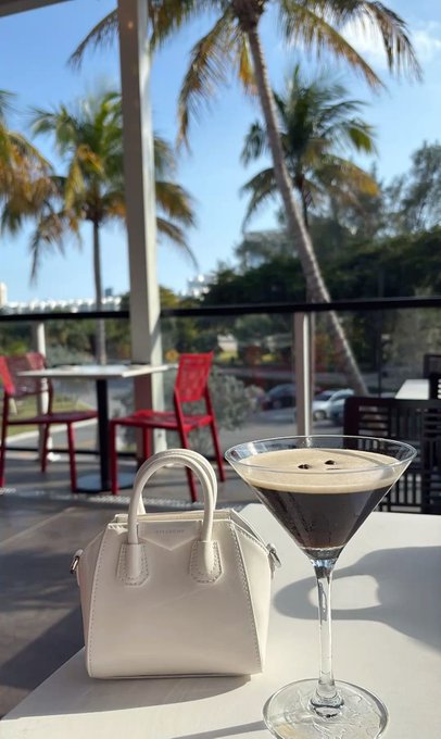 The image features a white purse sitting next to a martini glass filled with an interesting drink. The purse, which appears to be made of leather or fabric, has a small amount of liquid inside it. The drink itself seems to have a unique combination of ingredients that make it visually appealing and intriguing. The scene takes place in front of a patio where there are several chairs and dining tables arranged for people to enjoy their time outdoors.