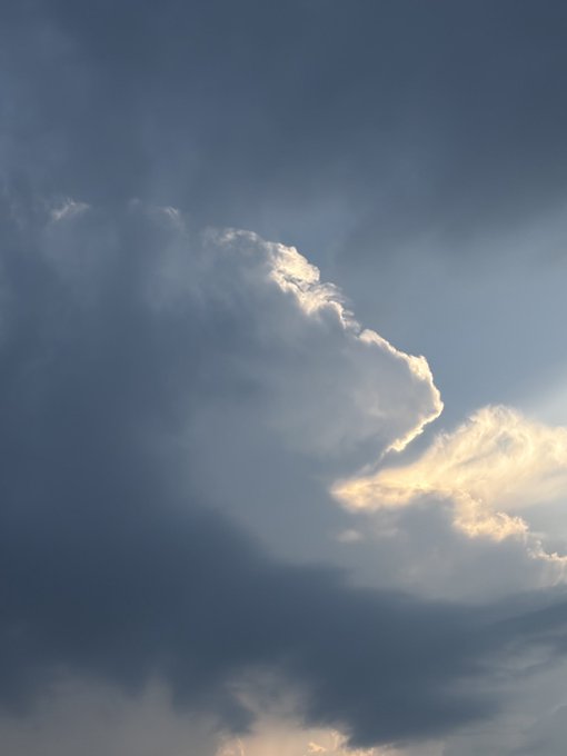 A dramatic sky scene featuring a large cumulus cloud backlit by the sun, with sun rays piercing through the cloud edges against a deep blue sky.
