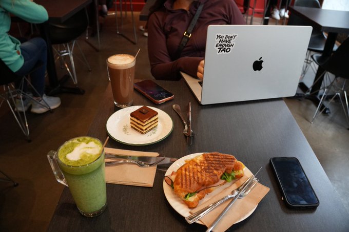 A café table scene featuring a matcha latte, iced coffee, a green tea tiramisu, and a pressed sandwich alongside a MacBook with a "YOU DON'T KNOW ENOUGH $TAO" decal—reinforcing the post's playful jab that the user is underinformed about Bittensor ($TAO).