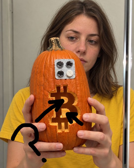 A woman in a yellow shirt holds a carved pumpkin decorated with a Bitcoin symbol and a blister pack of pills, with a black arrow and question mark drawn on the image.