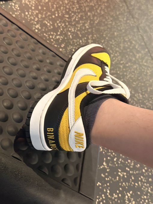 A person's foot wearing a yellow, black, and white Nike sneaker with 'BINARY' text on the side, photographed on a textured metal floor.